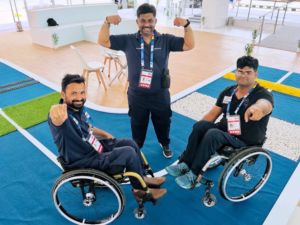 Mohammad Imran pointing forward alongside a standing teammate flexing biceps and fellow wheelchair user on the mobility obstacle track